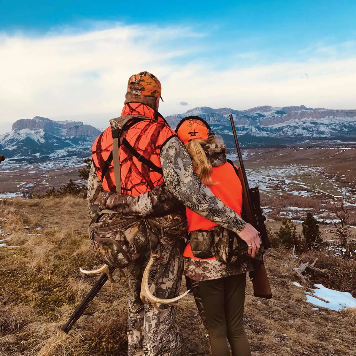 Two hunters in blaze orange looking out over a valley toward snowy mountains